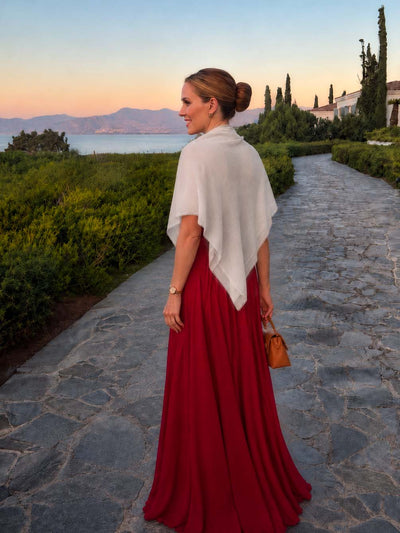 Woman wearing an ivory Square Featherlight Cashmere Scarf draped over her shoulders with an elegant red evening gown, walking along a coastal garden path at sunset.