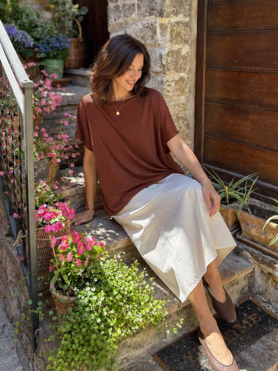Woman sitting on stone steps wearing cocoa silk top surrounded by flowers and plants