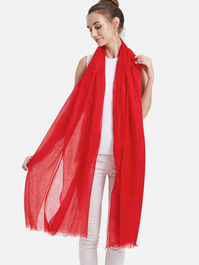A woman modeling a red featherlight cashmere scarf draped over her shoulders, paired with a white sleeveless top and white pants, against a solid light gray background.