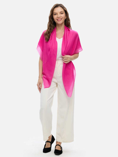 A woman modeling a fuchsia cashmere scarf draped around her neck, paired with a white sleeveless top and cream-colored trousers, against a solid light gray background.