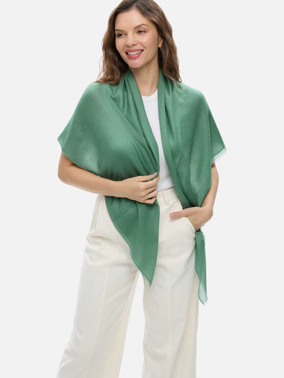A woman wearing a sage green cashmere scarf draped over her shoulders, paired with a white top and cream-colored trousers, against a solid light gray background.