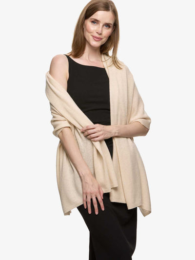 A woman modeling a light beige knit cashmere wrap or shawl draped over a black sleeveless top and black pants, photographed against a plain light gray studio background.