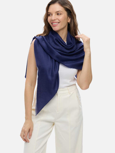 A woman modeling a navy blue Featherlight Square Cashmere Scarf wrapped around her shoulders, paired with a white sleeveless top and cream-colored trousers, against a solid light gray background.