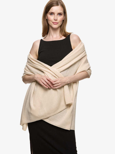 A woman modeling a light beige cashmere draped over a black off-the-shoulder top and black skirt, set against a plain light gray studio background.