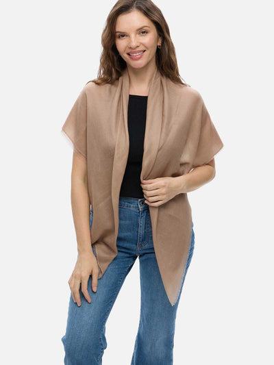 A woman modeling a camel Featherlight Square Cashmere Scarf draped over her shoulders, styled with a black top and blue jeans against a plain light gray studio background.