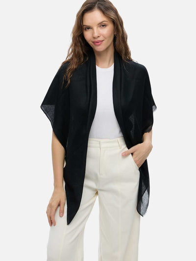 A woman modeling a black open-front shawl or lightweight cashmere scarf with sheer side panels, worn over a white top and cream trousers, against a plain light gray studio background.