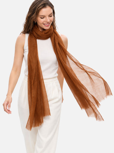 A woman wearing a long, semi-sheer featherlight chocolate cashmere scarf draped over her shoulders, paired with a white sleeveless top and white trousers, set against a plain light gray background.