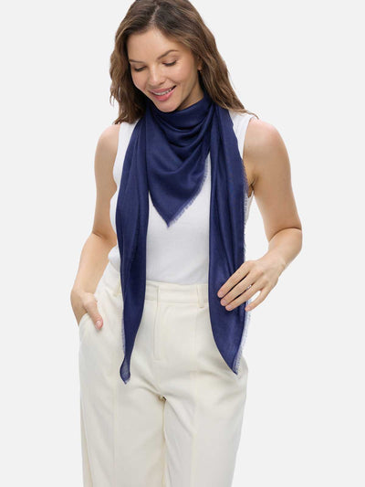 A woman modeling a navy blue Featherlight Square Cashmere Scarf wrapped around her neck over a white sleeveless top and cream-colored pants, set against a solid light gray background.