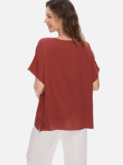 Woman wearing a rust-colored silk top and white pants on a light gray background