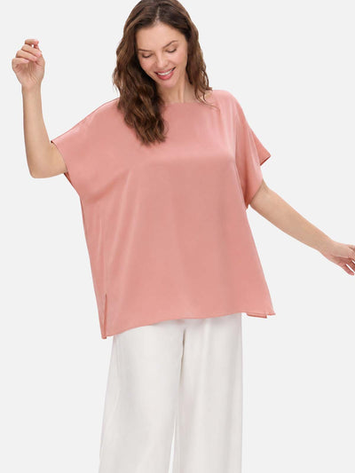 A woman wearing a loose, short-sleeve dusty pink silk top and white wide-leg pants, posing against a light gray studio background.