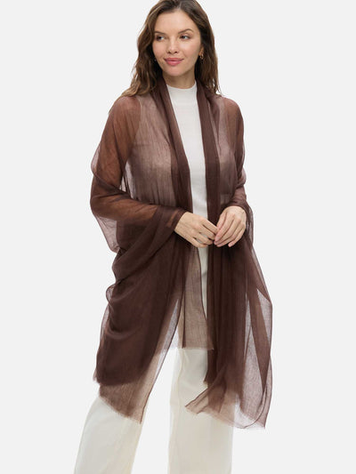 A woman modeling a sheer, lightweight featherlight brown cashmere scarf draped over a white top and white pants, set against a plain light gray studio background.