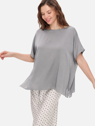 A woman wearing a loose, short-sleeve gray silk top and white pants with black polka dots, photographed against a plain light gray studio background.
