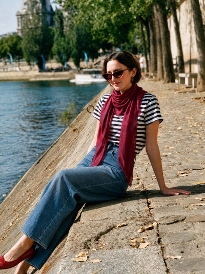 Woman wearing a wine-red Ovcio Featherlight Cashmere Scarf with a striped top and jeans by the river.