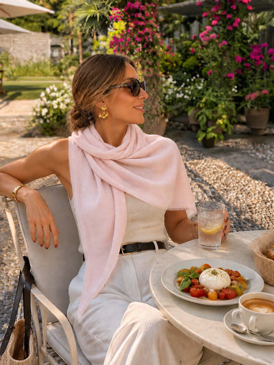 A woman sits at an outdoor patio table wearing a pink featherlight cashmere scarf draped over her shoulders, a white sleeveless top, and white pants. A plate of fresh burrata with tomatoes, a glass of water, and a cup of coffee are on the table in front of her.