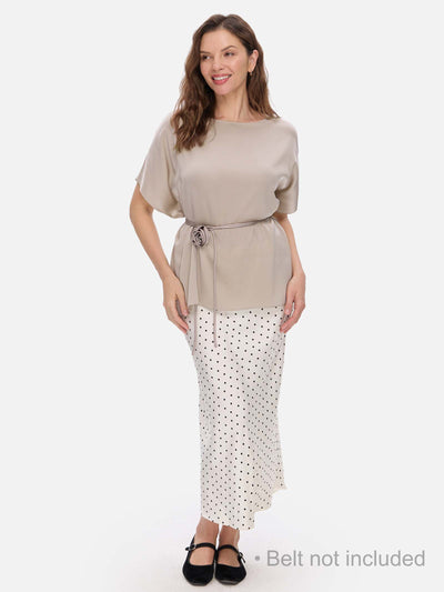 A woman modeling a two-piece outfit consisting of a beige short-sleeve silk top with a matching waist tie and a white midi skirt with black polka dots. She is wearing black flats against a solid light gray background.