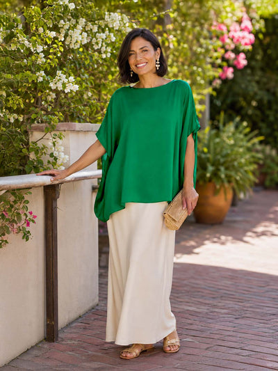 Woman in a green silk top and beige pants standing outdoors with greenery in the background