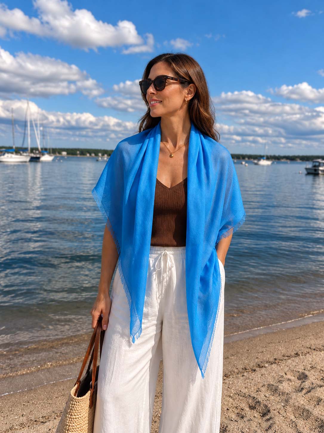 A woman wearing a bright blue Featherlight Square Cashmere Scarf draped over her shoulders, styled with sunglasses, a brown sleeveless top, and white wide-leg trousers by a sunny marina.