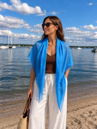 A woman wearing a bright blue Featherlight Square Cashmere Scarf draped over her shoulders, styled with sunglasses, a brown sleeveless top, and white wide-leg trousers by a sunny marina.