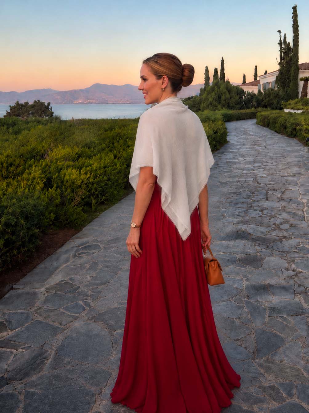 Woman wearing an ivory Square Featherlight Cashmere Scarf draped over her shoulders with an elegant red evening gown, walking along a coastal garden path at sunset.