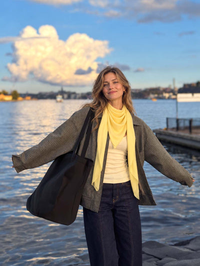 Woman wearing a yellow Featherlight Square Cashmere Scarf loosely wrapped around her neck, styled with a plaid jacket, white top, dark jeans, and a black tote by the waterfront at sunset.
