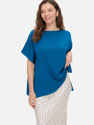 A woman modeling a bright blue silk top with a boat neckline and a white skirt with black polka dots, set against a solid light gray background.