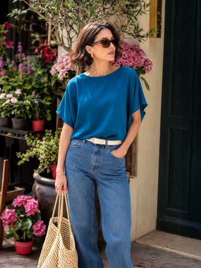 A woman wearing a vibrant teal blue short-sleeve silk top and blue jeans stands outdoors in front of a building with potted pink flowers. She is holding a woven straw tote bag.