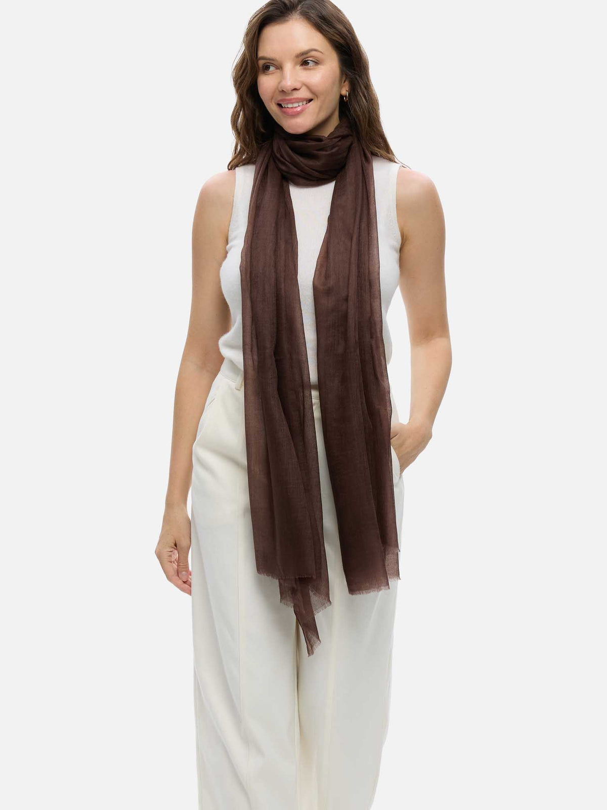 A woman modeling a long, semi-sheer featherlight brown cashmere scarf wrapped around her neck, paired with a white sleeveless top and white wide-leg pants, against a solid light gray background.