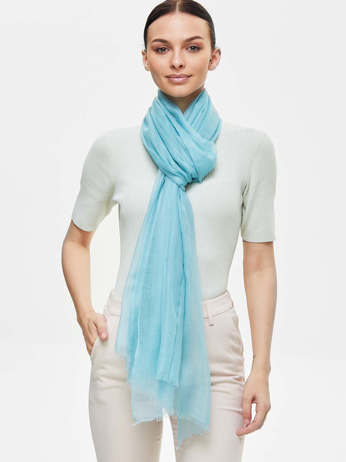 A woman modeling a featherlight light blue cashmere scarf wrapped around her neck, paired with a pale green ribbed short-sleeve top and beige trousers, against a plain white studio background.