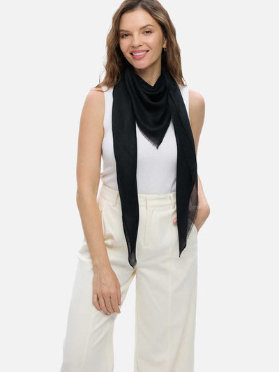 A woman modeling a lightweight black cashmere scarf draped around her neck over a white sleeveless top and cream-colored trousers, set against a plain light gray studio background.