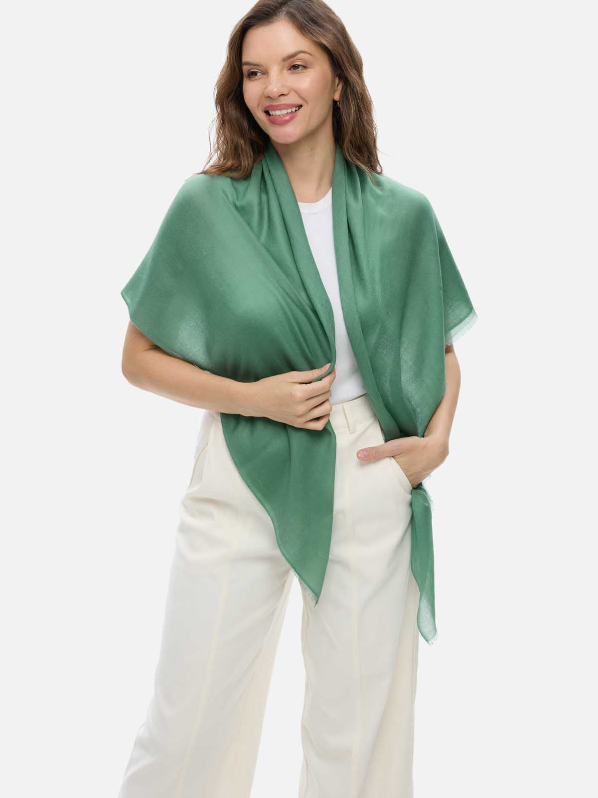 A woman wearing a sage green cashmere scarf draped over her shoulders, paired with a white top and cream-colored trousers, against a solid light gray background.