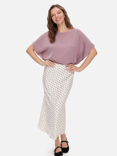 A woman modeling a mauve silk top and a white midi skirt with black polka dots, standing against a light gray studio background.