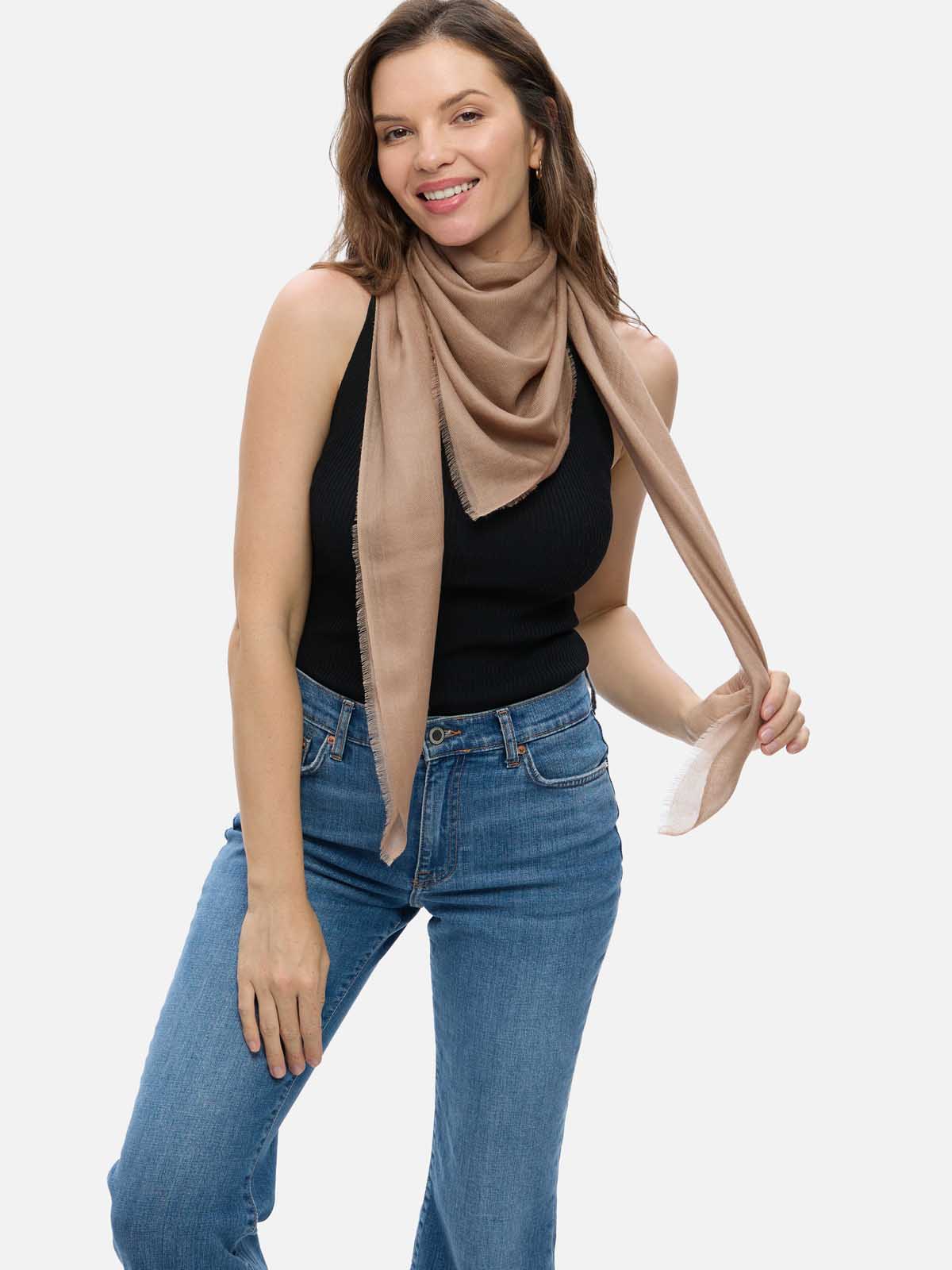 A woman modeling a camel Featherlight Square Cashmere Scarf draped over her shoulders, styled with a black top and blue jeans against a plain light gray studio background.