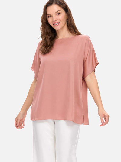 A woman wearing a dusty rose short-sleeve silk top and white pants, standing against a light gray studio background.