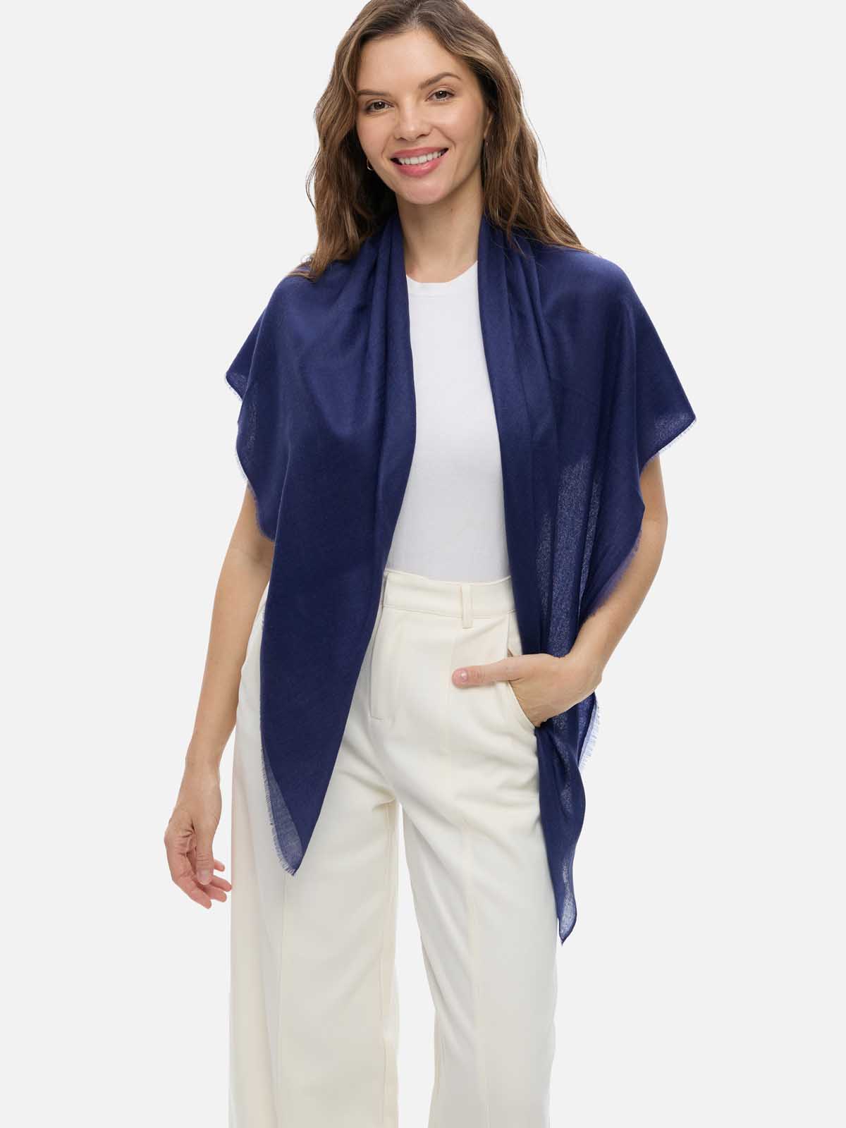 A woman modeling a navy blue Featherlight Square Cashmere Scarf draped over her shoulders, worn over a white top and white trousers, against a plain light gray studio background.