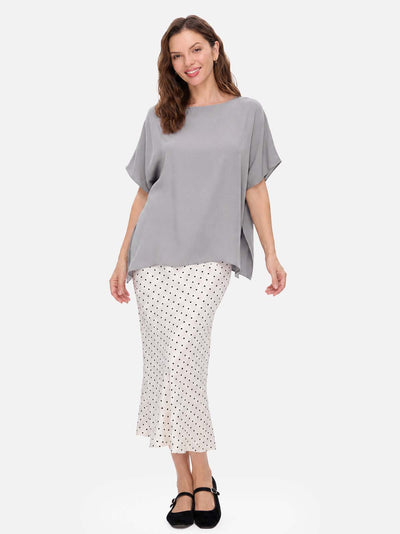 A woman modeling a loose-fit gray short-sleeve silk top paired with a white midi skirt featuring a black polka dot pattern, against a solid light gray background.