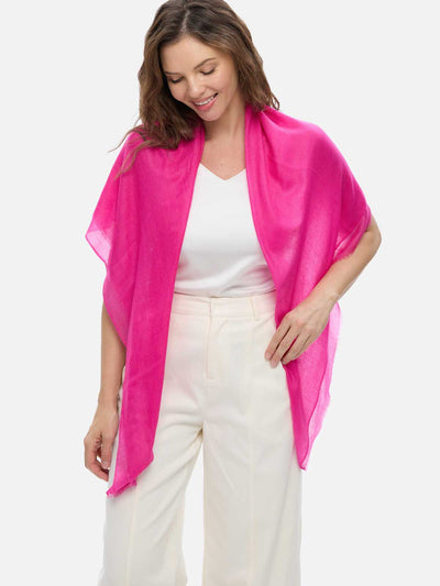 A woman modeling a fuchsia cashmere scarf draped around her neck, paired with a white sleeveless top and cream-colored trousers, against a solid light gray background.
