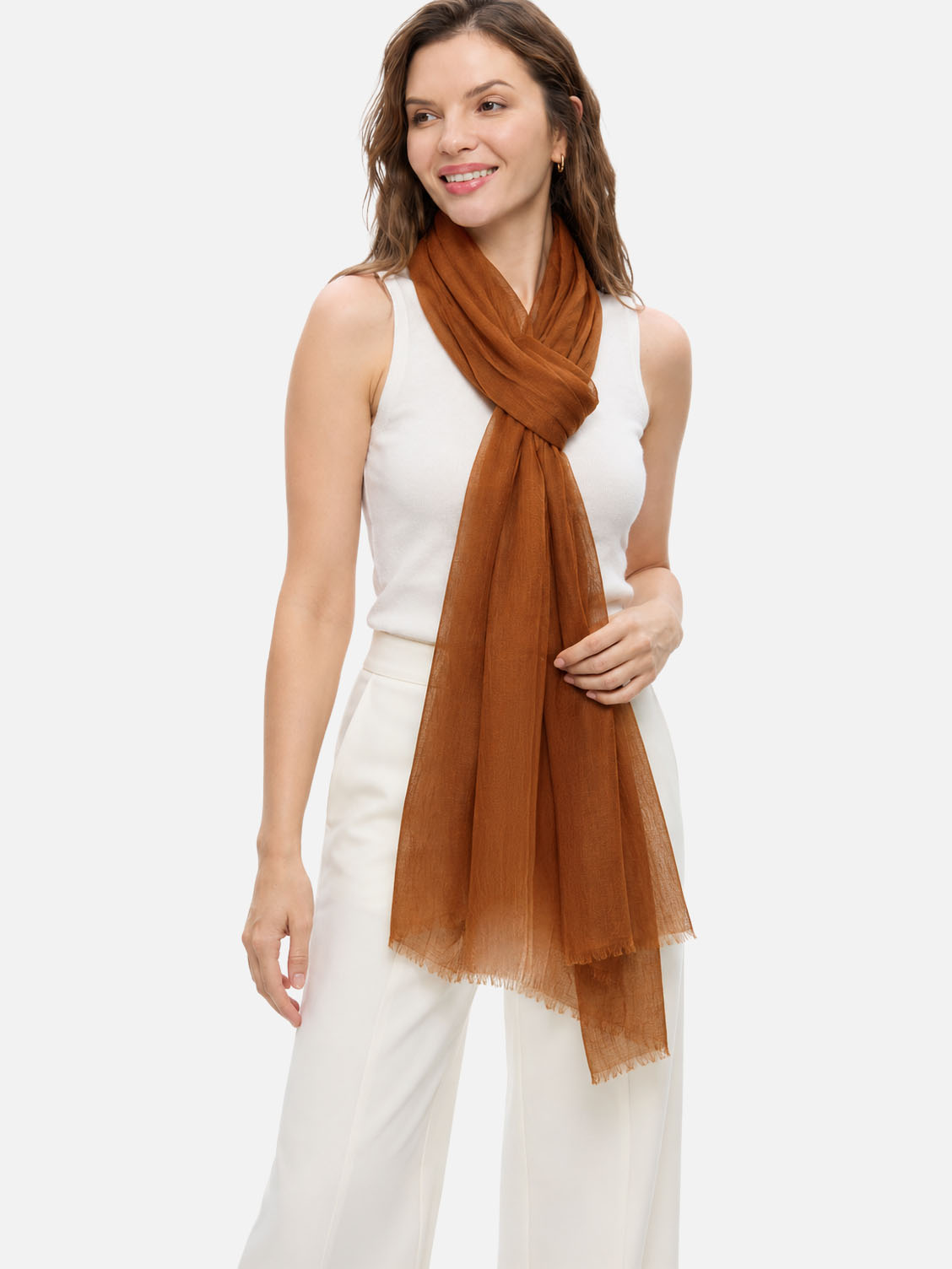 A woman modeling a lightweight, semi-sheer featherlight chocolate cashmere scarf in a warm rust brown color, styled over a white sleeveless top and white wide-leg trousers against a solid light gray background.