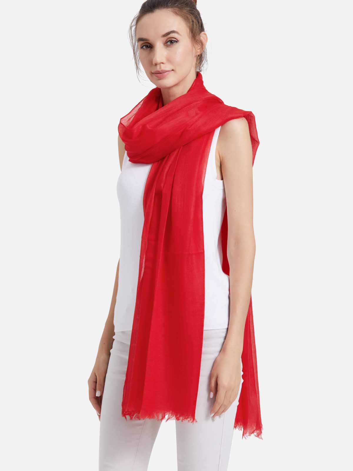 A woman modeling a red featherlight cashmere scarf wrapped around her shoulders and draped down her front, paired with a white sleeveless top and white pants against a light gray studio background.