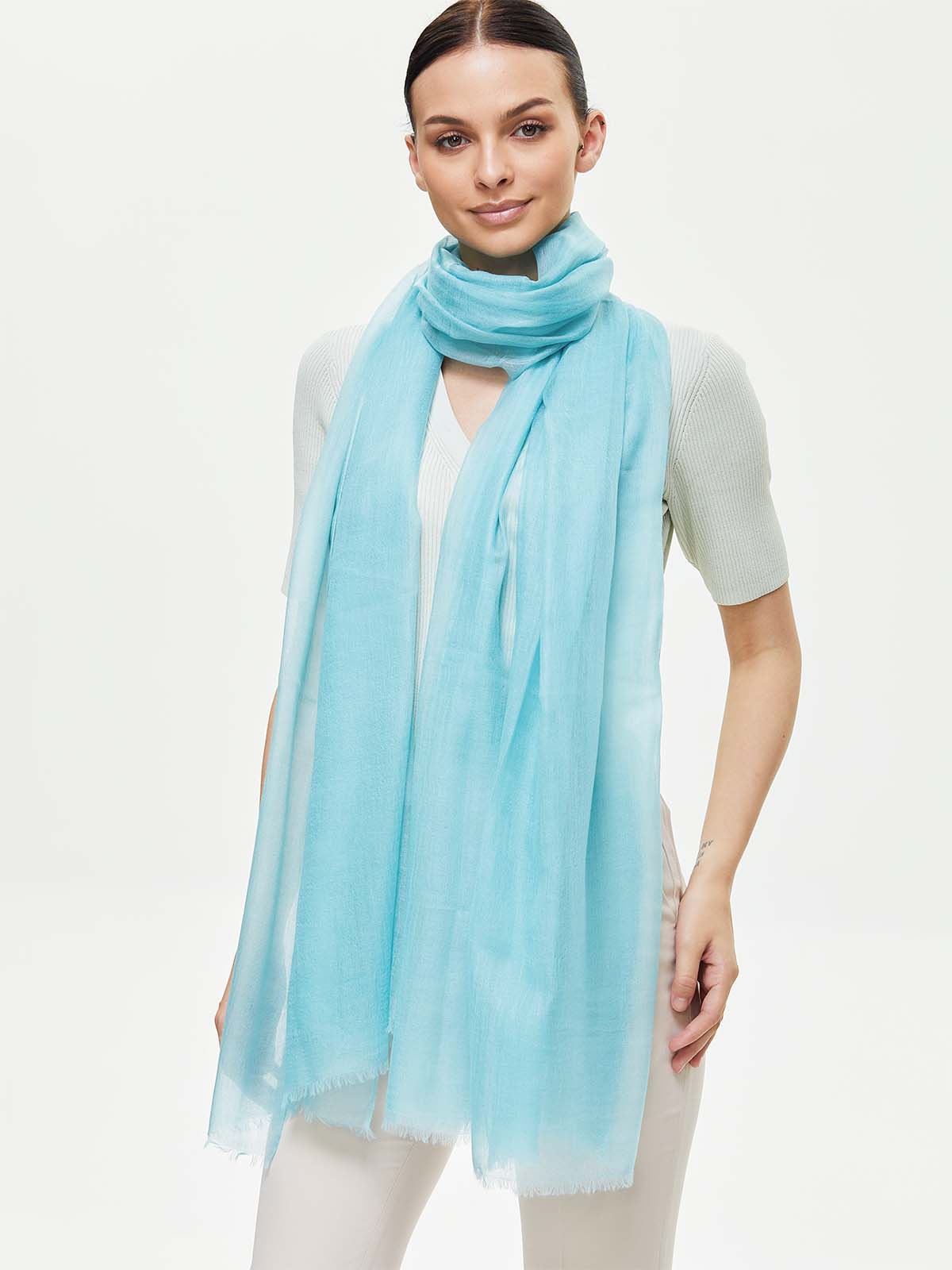 A woman modeling a featherlight light blue cashmere scarf draped over her shoulders, paired with a white short-sleeve top and white pants against a plain white studio background.