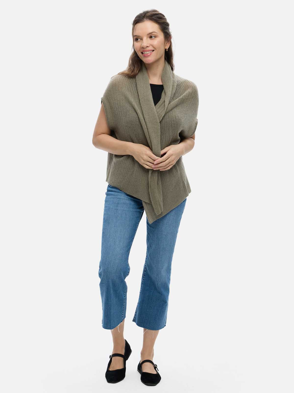 Woman wearing a green cashmere vest over a black top and blue jeans on a white background