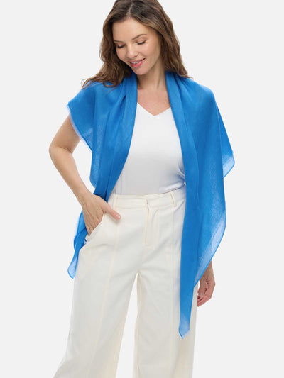 A woman wearing a blue Featherlight Square Cashmere Scarf draped over a white sleeveless top and cream-colored trousers, photographed against a light gray studio background.