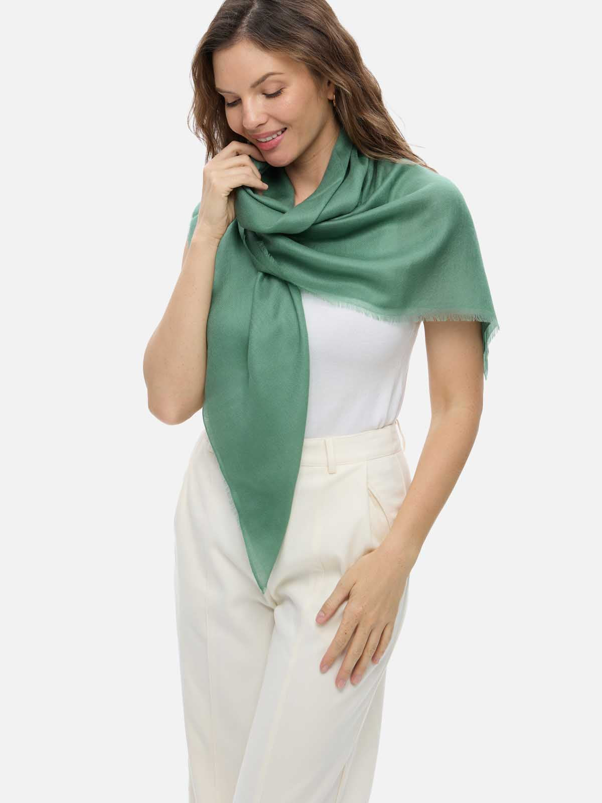 A woman wearing a sage green cashmere scarf draped over her shoulders, paired with a white top and cream-colored trousers, against a solid light gray background.