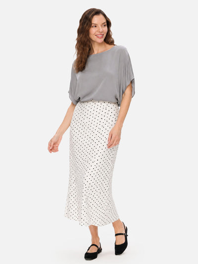 A woman modeling a gray short-sleeve silk top and a white midi skirt with black polka dots, paired with black Mary Jane shoes, against a light gray studio background.
