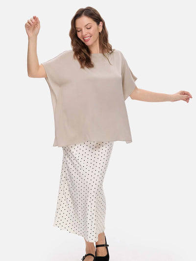 A woman modeling a loose, short-sleeve taupe silk top paired with a white skirt featuring a black polka-dot pattern, set against a plain light gray studio background.