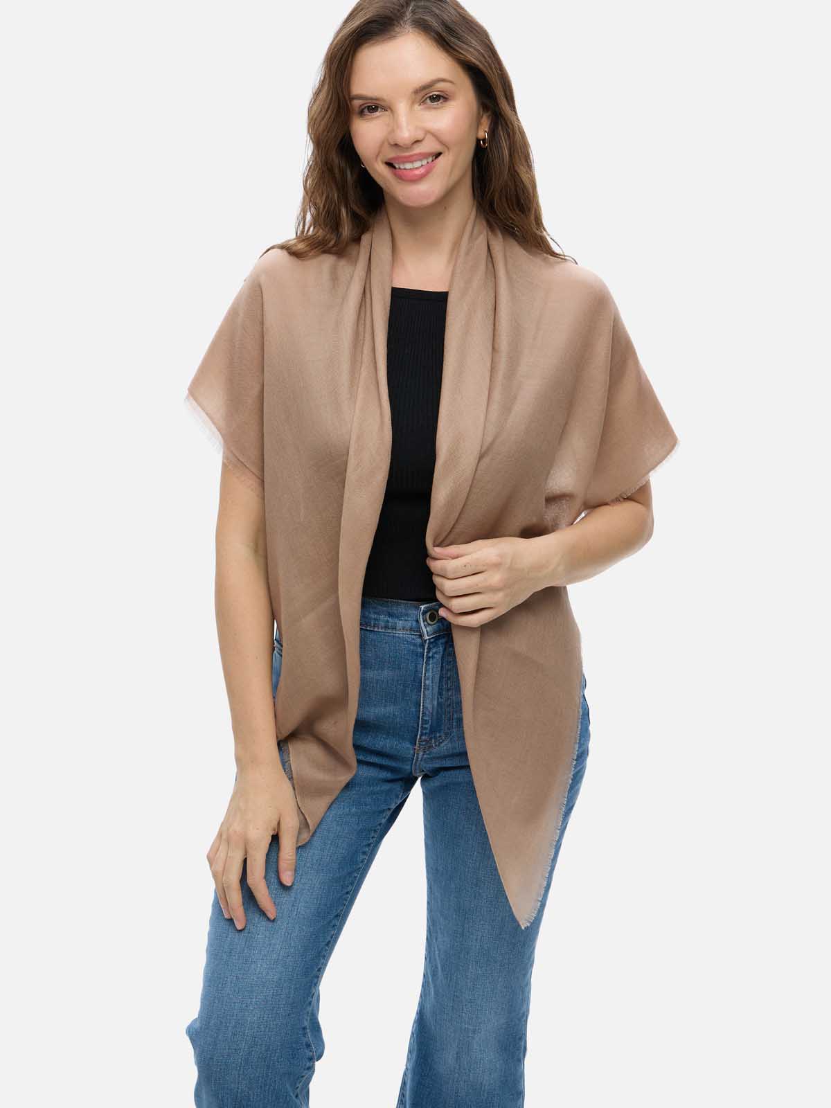 A woman modeling a camel Featherlight Square Cashmere Scarf draped over her shoulders, styled with a black top and blue jeans against a plain light gray studio background.