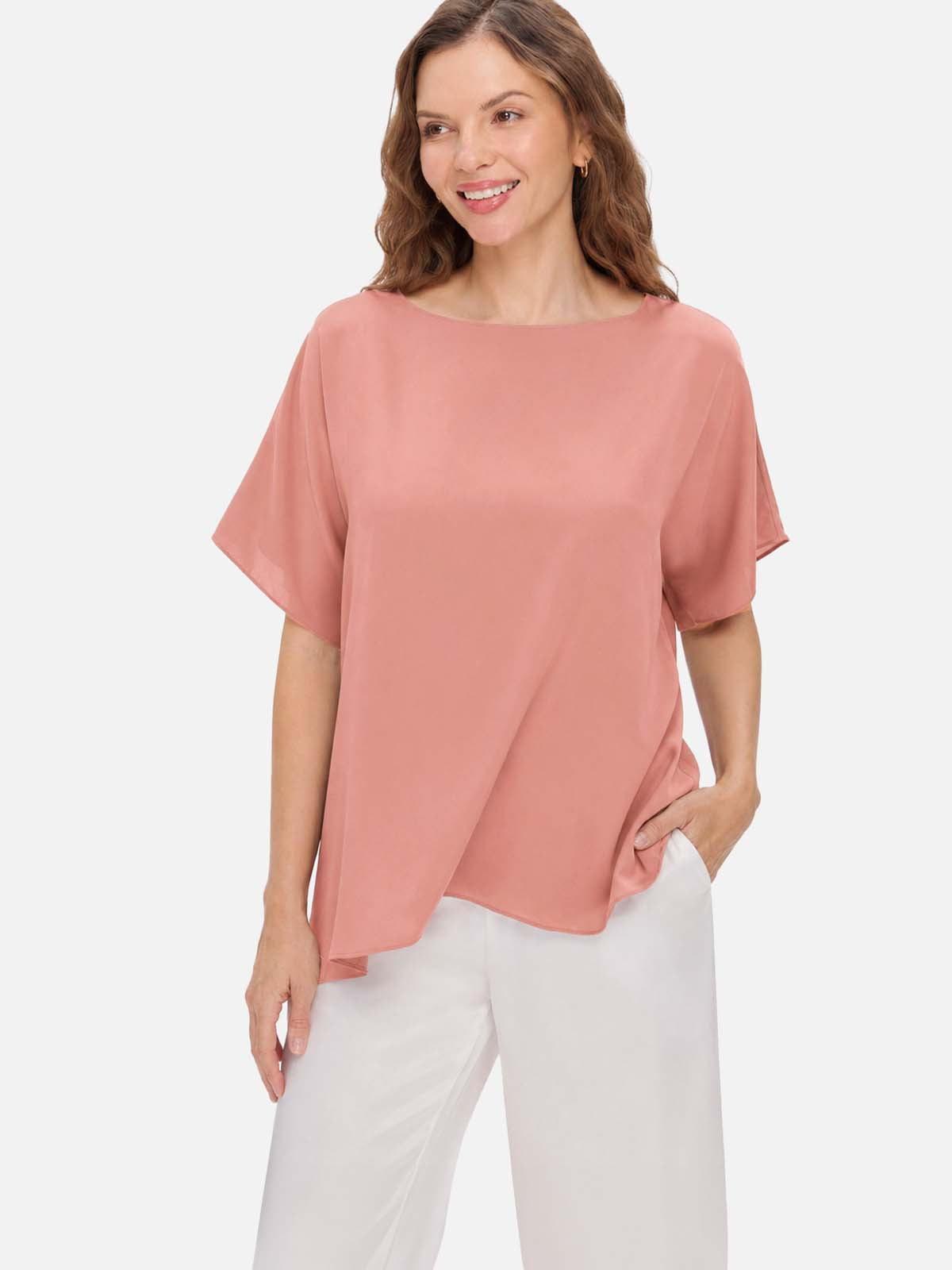 A woman wearing a loose, short-sleeve dusty rose silk top and white pants, standing against a light gray studio background.