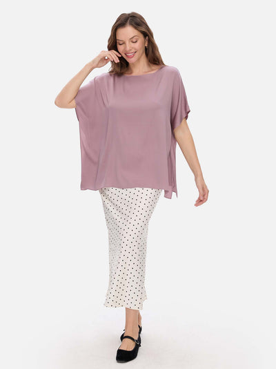 A woman modeling a loose, short-sleeve mauve silk top paired with white pants featuring a black polka-dot pattern and black Mary Jane shoes, set against a plain light gray background.