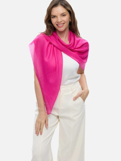 A woman modeling a fuchsia cashmere scarf draped around her neck, paired with a white sleeveless top and cream-colored trousers, against a solid light gray background.