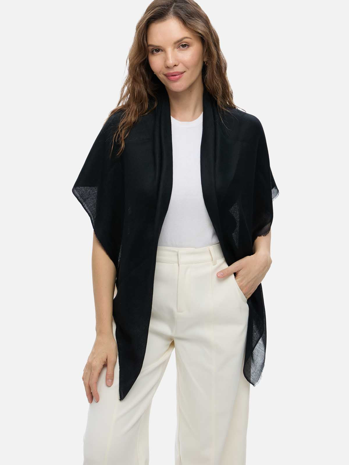 A woman modeling a black open-front shawl or lightweight cashmere scarf with sheer side panels, worn over a white top and cream trousers, against a plain light gray studio background.