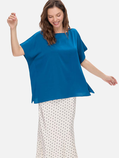 A woman modeling a bright blue short-sleeve silk top with a boat neckline and flared sleeves, paired with a white skirt with black polka dots, against a light gray studio background.